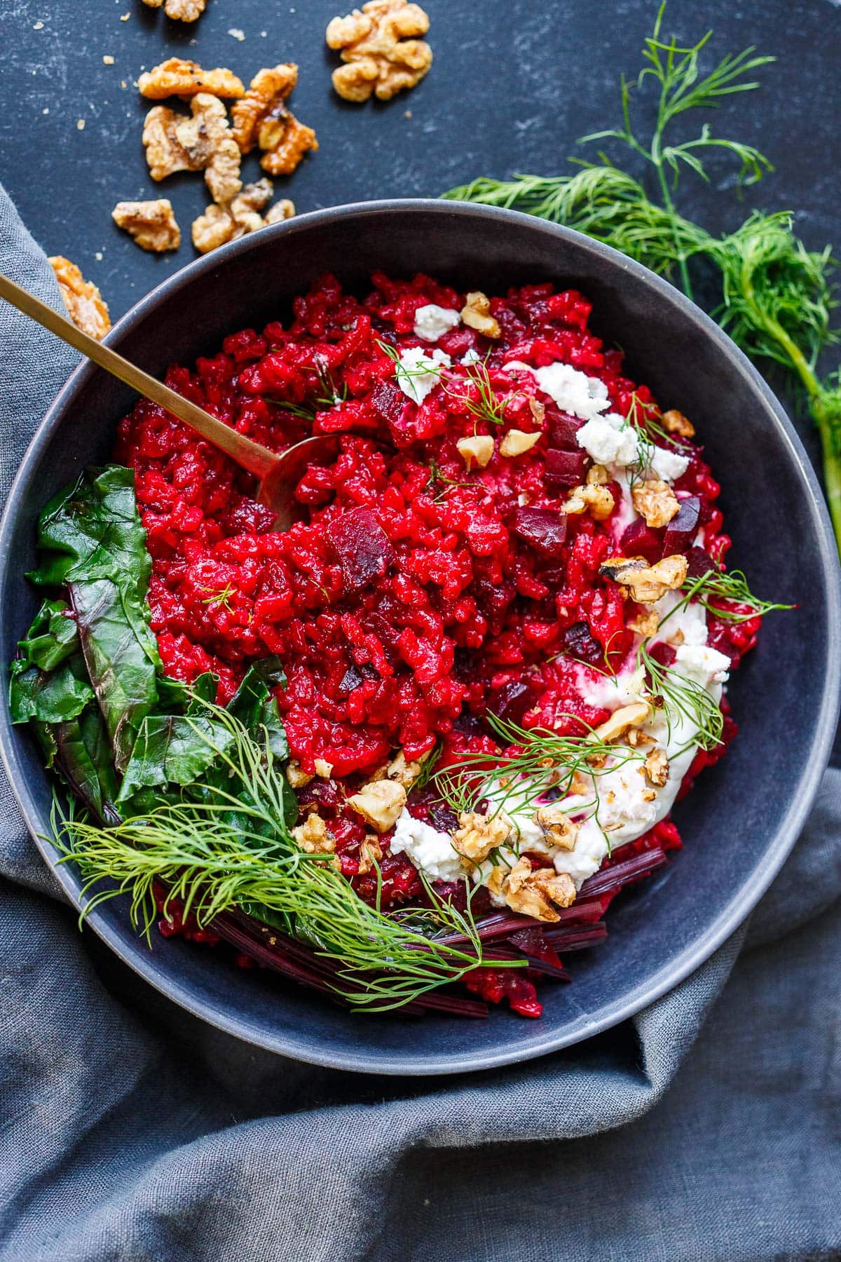 bowl of beet risotto with spoon, topped with goat cheese, toasted walnuts, fresh dill, and steamed beet greens.