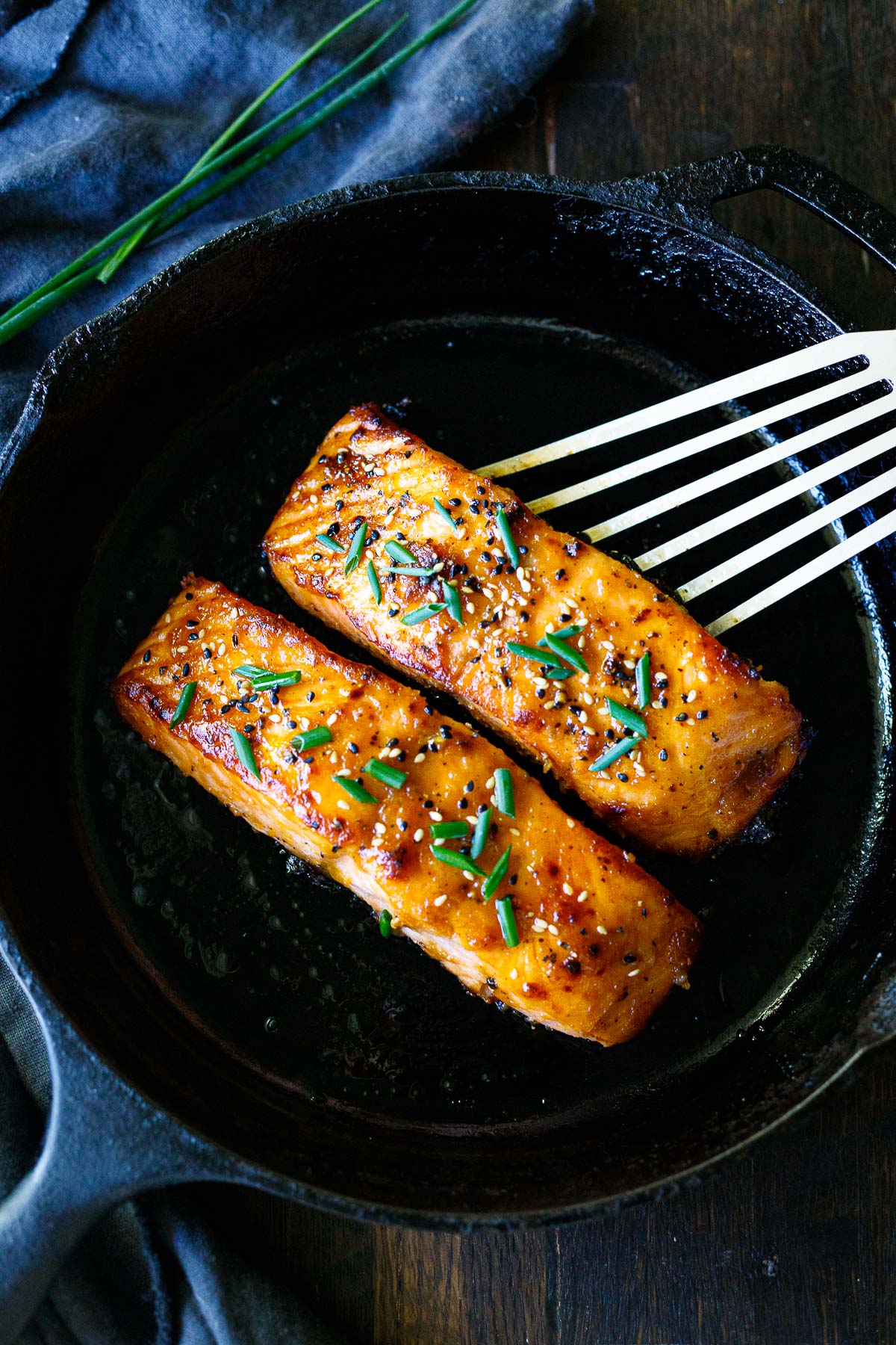 two broiled salmon filets on a cast iron skillet with sesame seeds and chives as garnish with a metal spatula beginning to lift one up.
