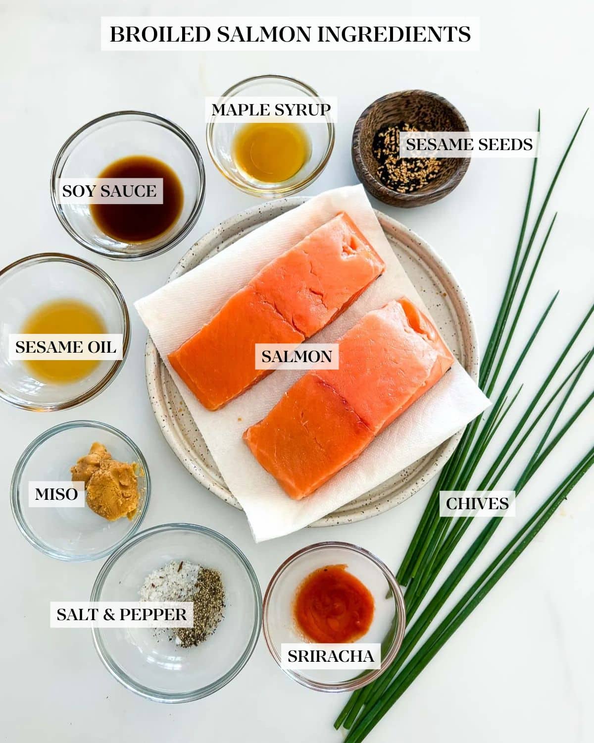 ingredients laid out for broiled salmon - two salmon filets on a plate, surrounded by small bowls of sesame seeds, sesame oil, soy sauce, olive oil, miso paste, salt and pepper, and sriracha, next to chives.