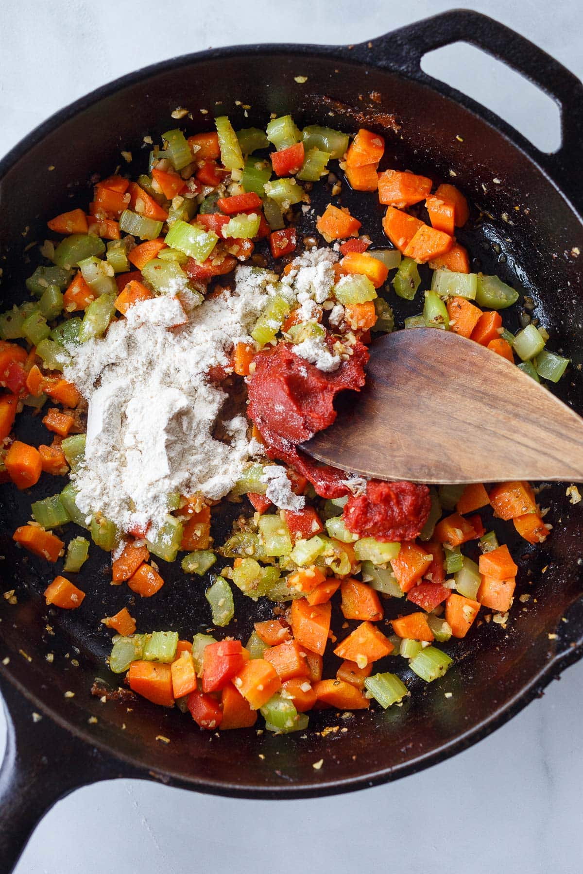 wooden spoon stirring carrots, celery, tomato paste, and flour in cast iron skillet. 