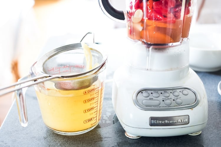 measuring cup with brine with a small strainer on top next to a blender with the fermented chilies and some brine.