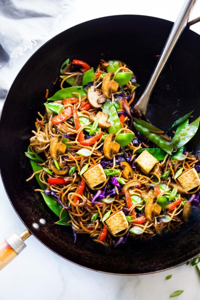 veggie lo mein noodles in a wok with cubed tofu, snow peas, mushrooms, peppers, green onions, and cabbage.