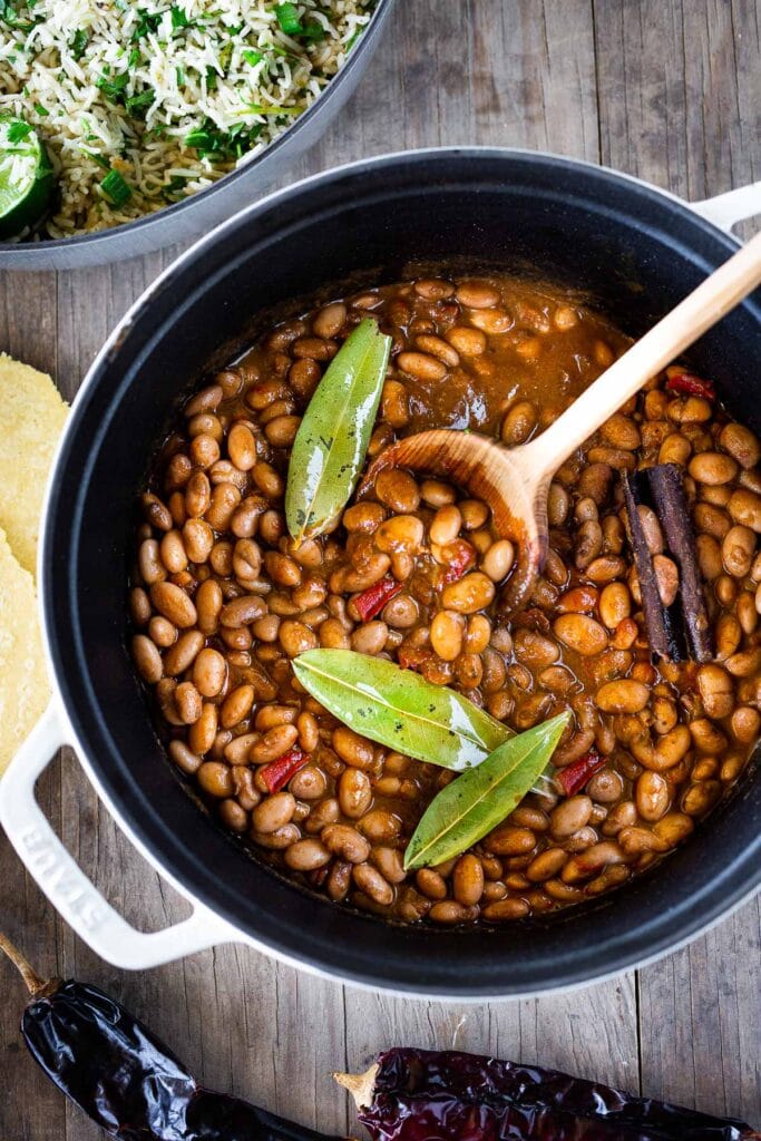 large pot with Mexican pinto beans with cinnamon stick, bay leaves, and. awood spoon for serving.