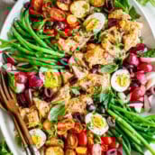 nicoise salad with salmon, boiled eggs, roasted potatoes, green beans, olives, radishes, tomatoes, and herbs on serving platter with utensil, one of our Mediterranean recipes.