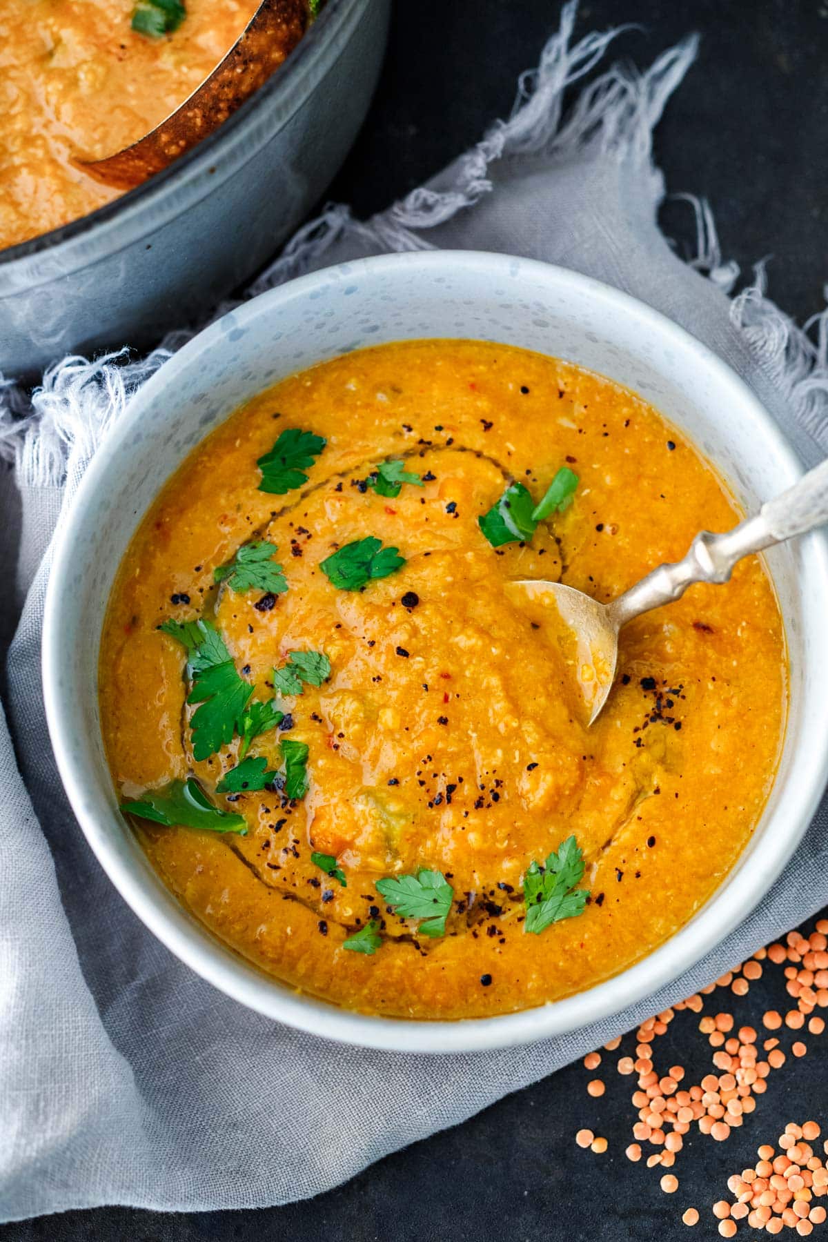 bowl of red lentil woup with spoon, garnished with swirl of oil, spices, and fresh herbs.