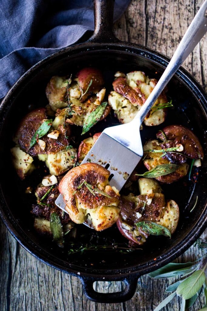 cast iron skillet with metal spatula holding up an herby smashed potato with more on the skillet - topped with herbs and garlic.