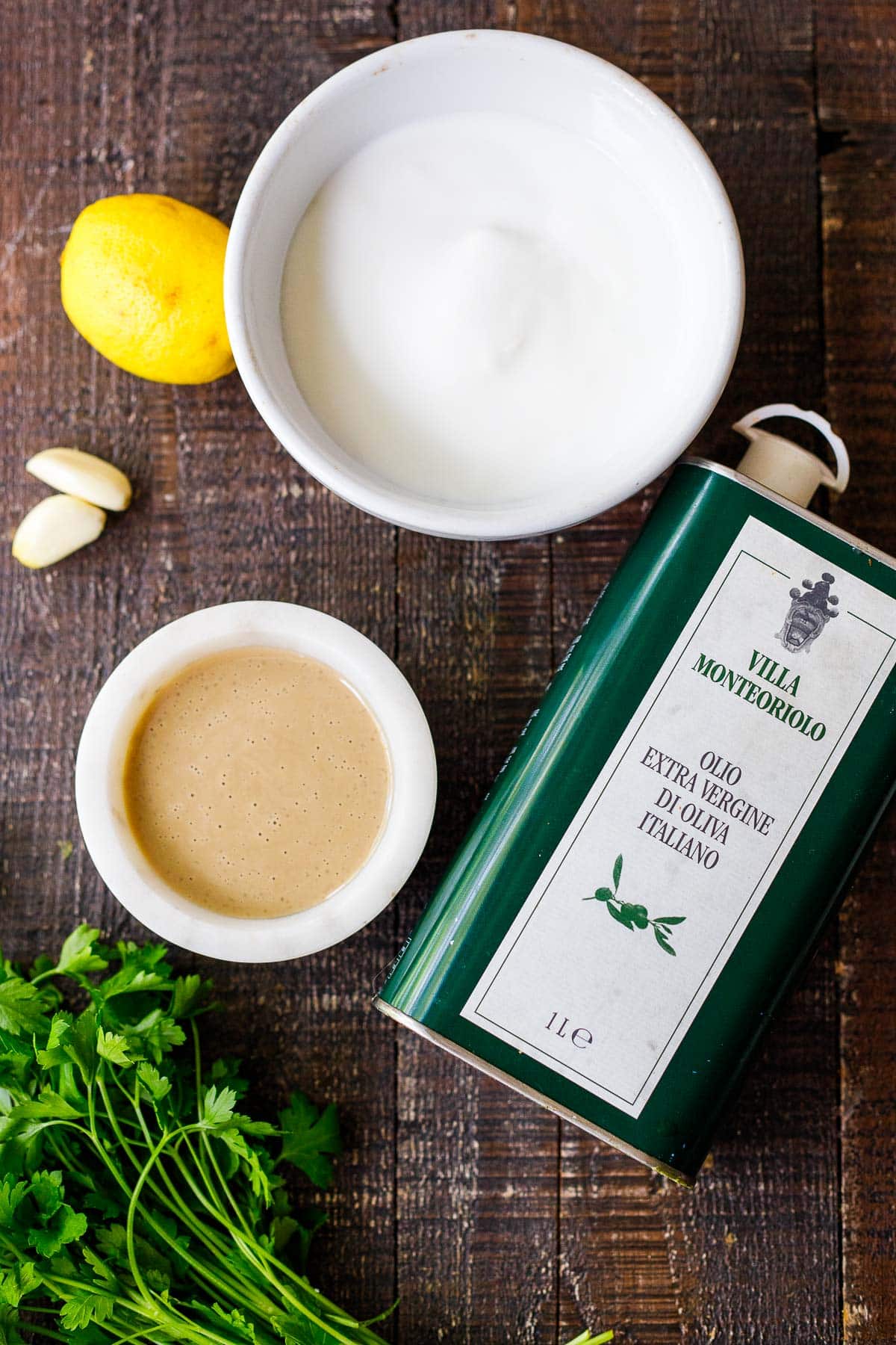 ingredients for tahini yogurt sauce - bowl of greek yogurt, tahini, olive oil, garlic cloves, lemon, parsley. 