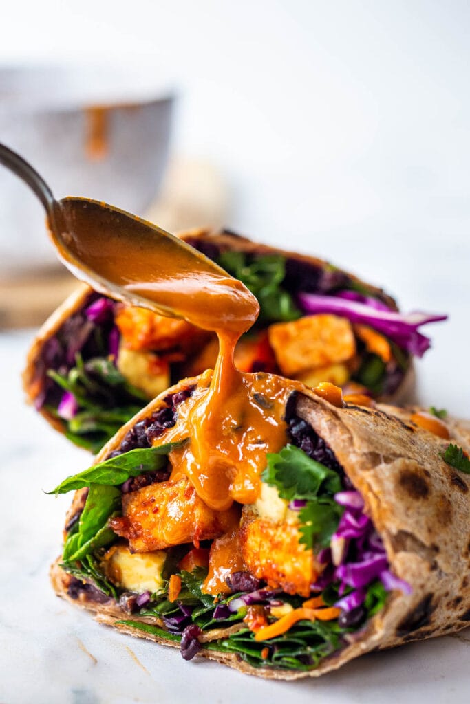 Thai burrito with tofu, black rice, purple cabbage, and herbs with a spoon drizzling peanut sauce over top.