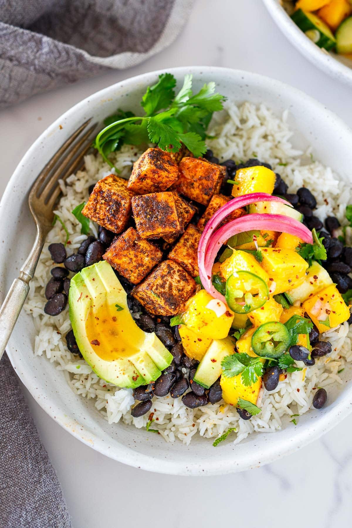 spicy jerk tofu bowl over rice and black beans, with avocado, mango, jalapeño, cilantro, pickled onions, and jerk tofu.