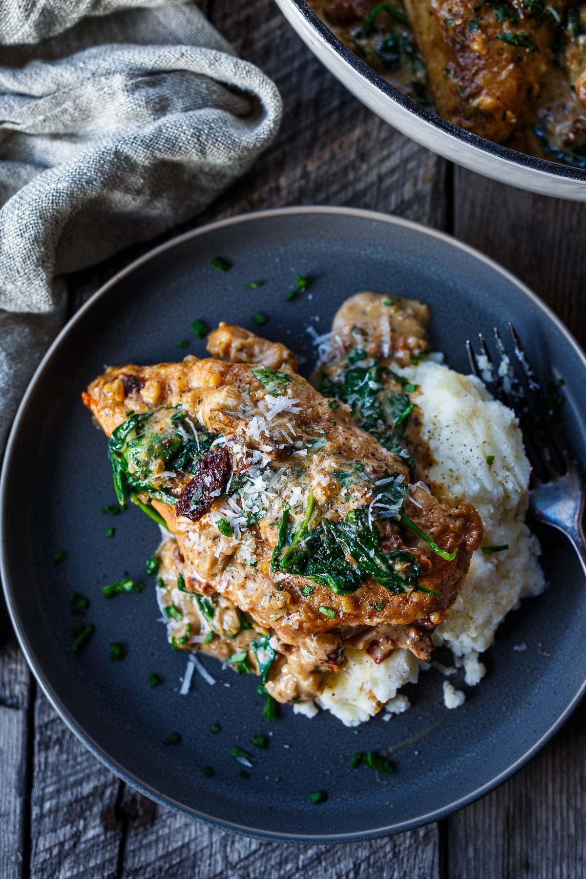 plate of creamy Tuscan chicken on mashed potatoes with fork, topped with spinach and sun-dried tomatoes and pecorino. 