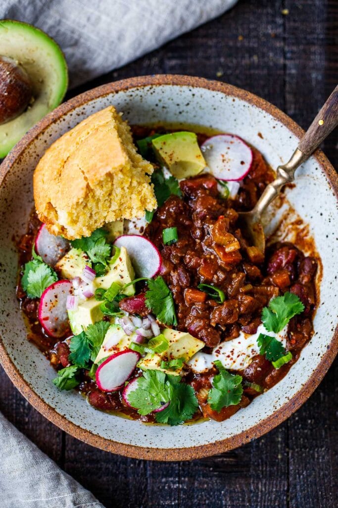 vegan chili topped with radish, avocado, red onion, green onion, cilantro, and vegan sour cream with a piece of cornbread in a bowl with spoon.