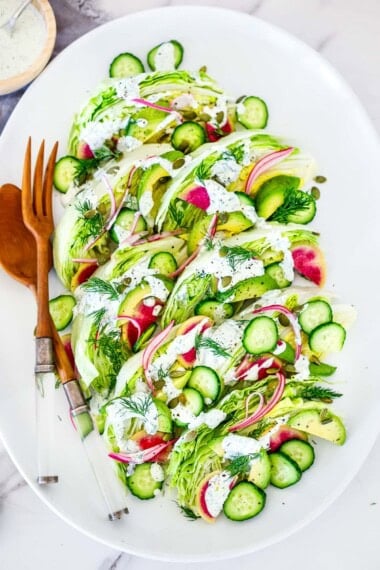 oval platter with wedge salad with red onion, cucumber, avocado, pepitas, watermelon radish, and fresh dill, topped with creamy dressing, served with wood utensils.